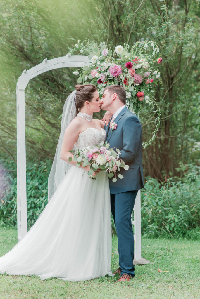 Bride and Groom standing under white lattice arbor decorated with pink, burgundy and lavender colored florals. Shows the story of a wedding day.
